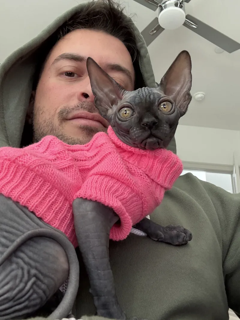 Eddy holding Tini, his black Sphynx cat, in a pink sweater